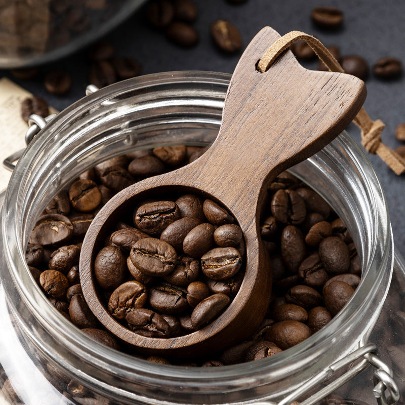 Wooden scoop filled with coffee beans inside a glass jar, with scattered coffee beans around.