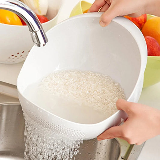 Person washing rice in a white colander over a sink with fruits in the background